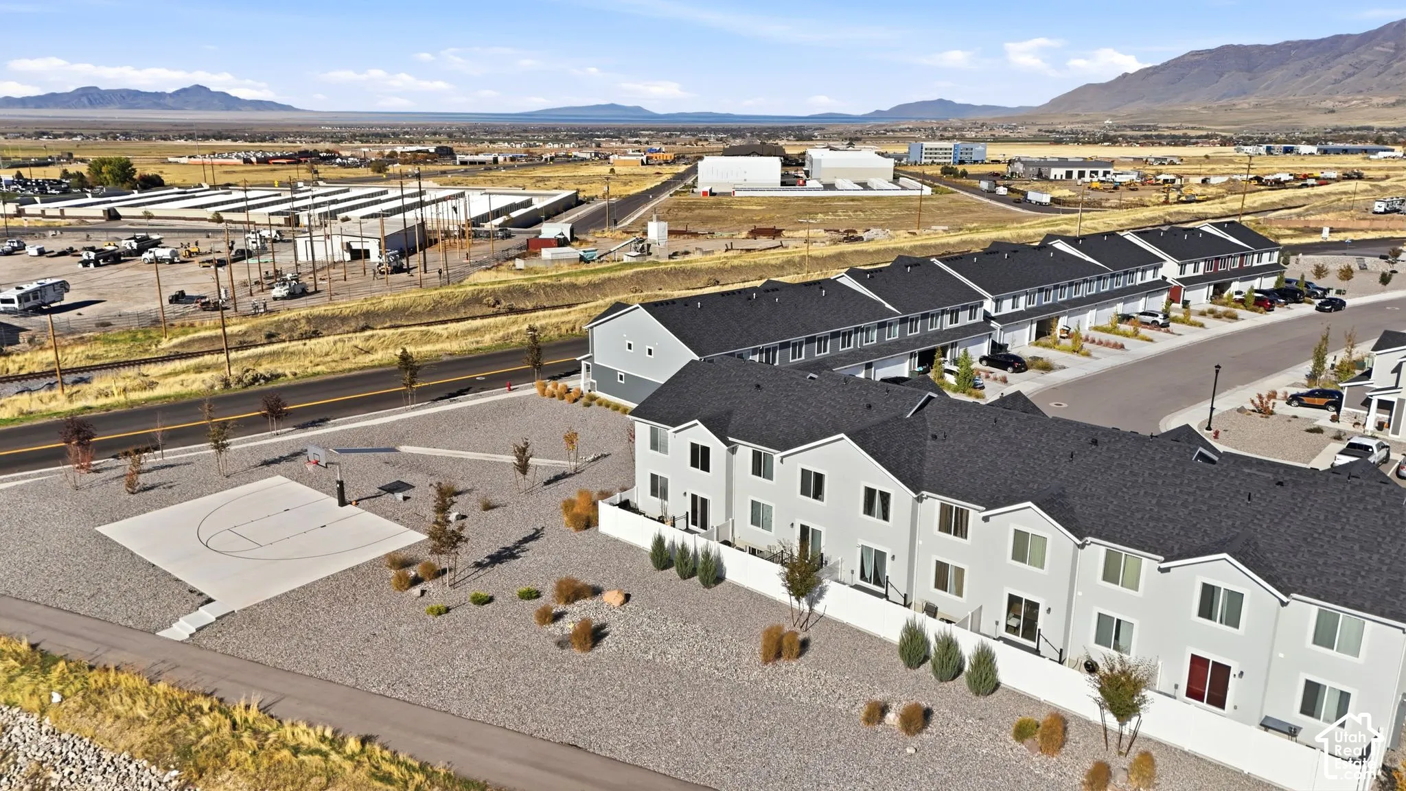Multi-level townhome featuring a dark shingle roof, neutral exterior siding, a concrete basketball court, and xeriscaped grounds with mountain backdrops