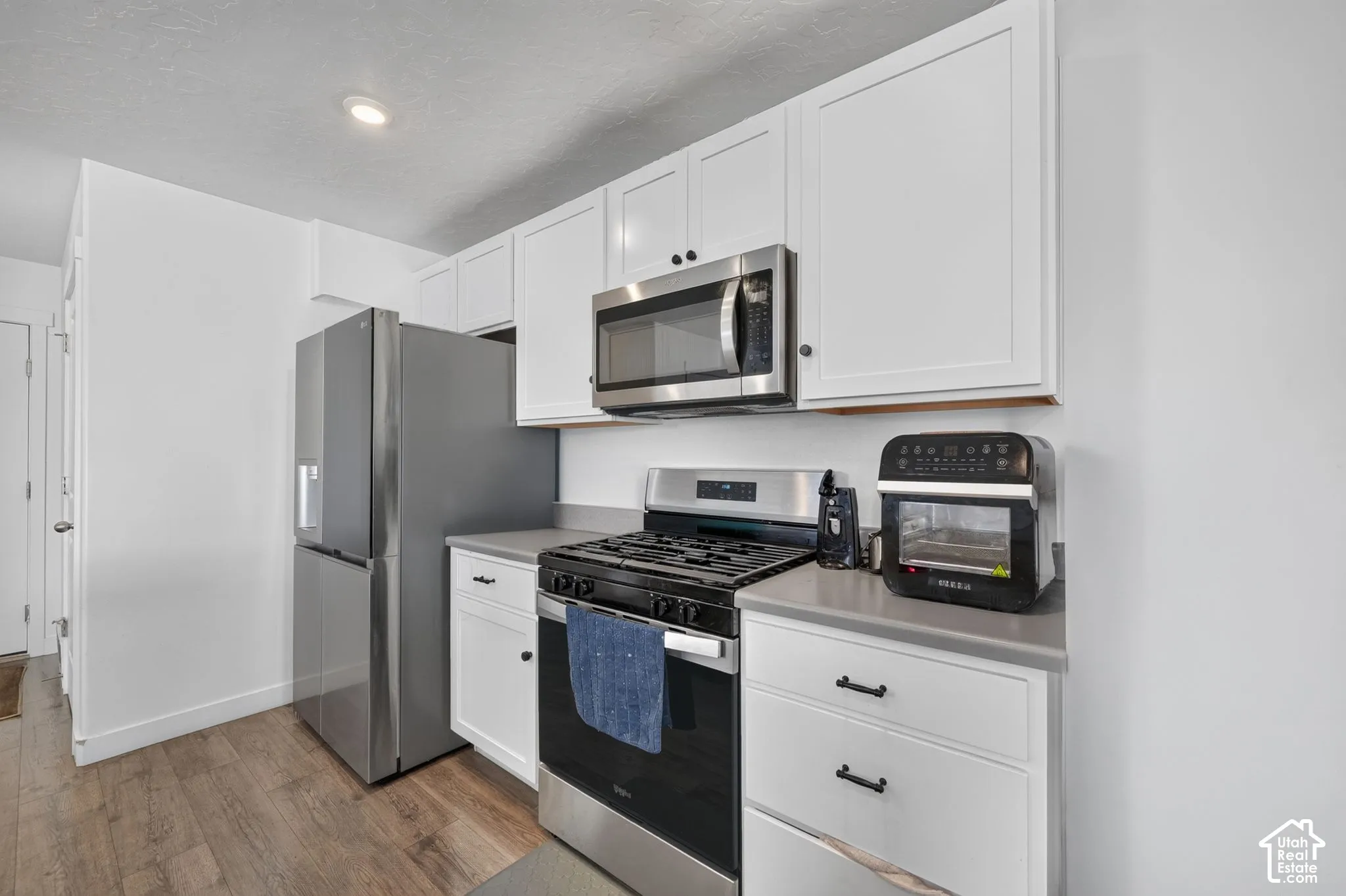 Kitchen featuring white shaker-style cabinetry, solid-surface countertops, wood-finish flooring, and stainless steel appliances including a gas range and microwave