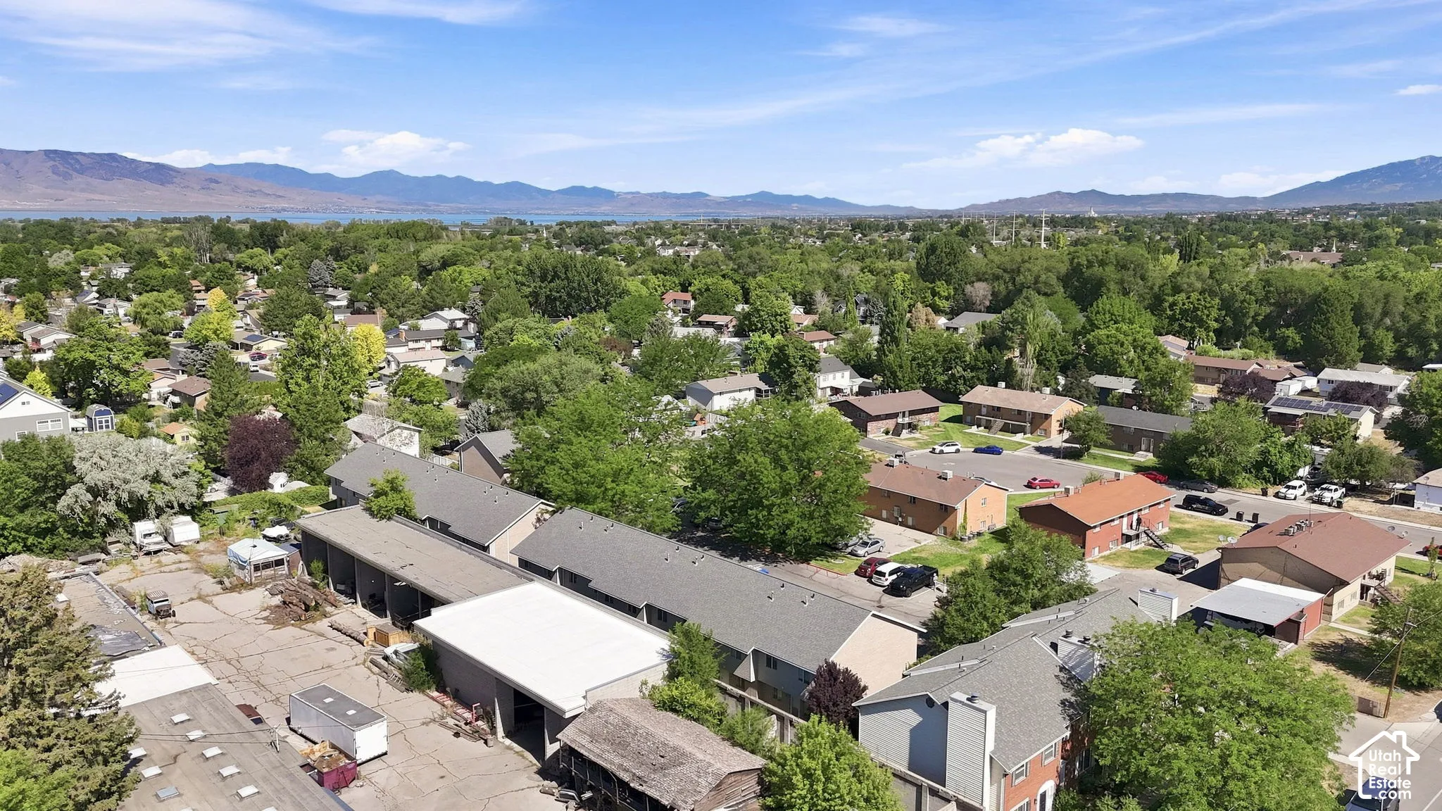 Aerial perspective of the residential area featuring multiple low-rise structures, paved parking surfaces, mature trees, and a distant mountain backdrop