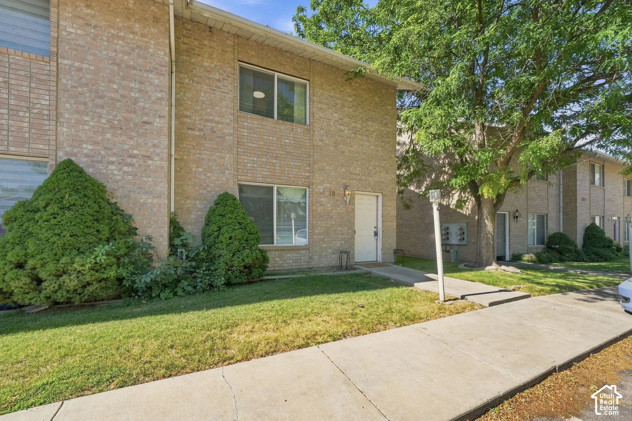Multi-story tan brick exterior featuring sliding windows, mature trees, manicured landscaping, and concrete walkways