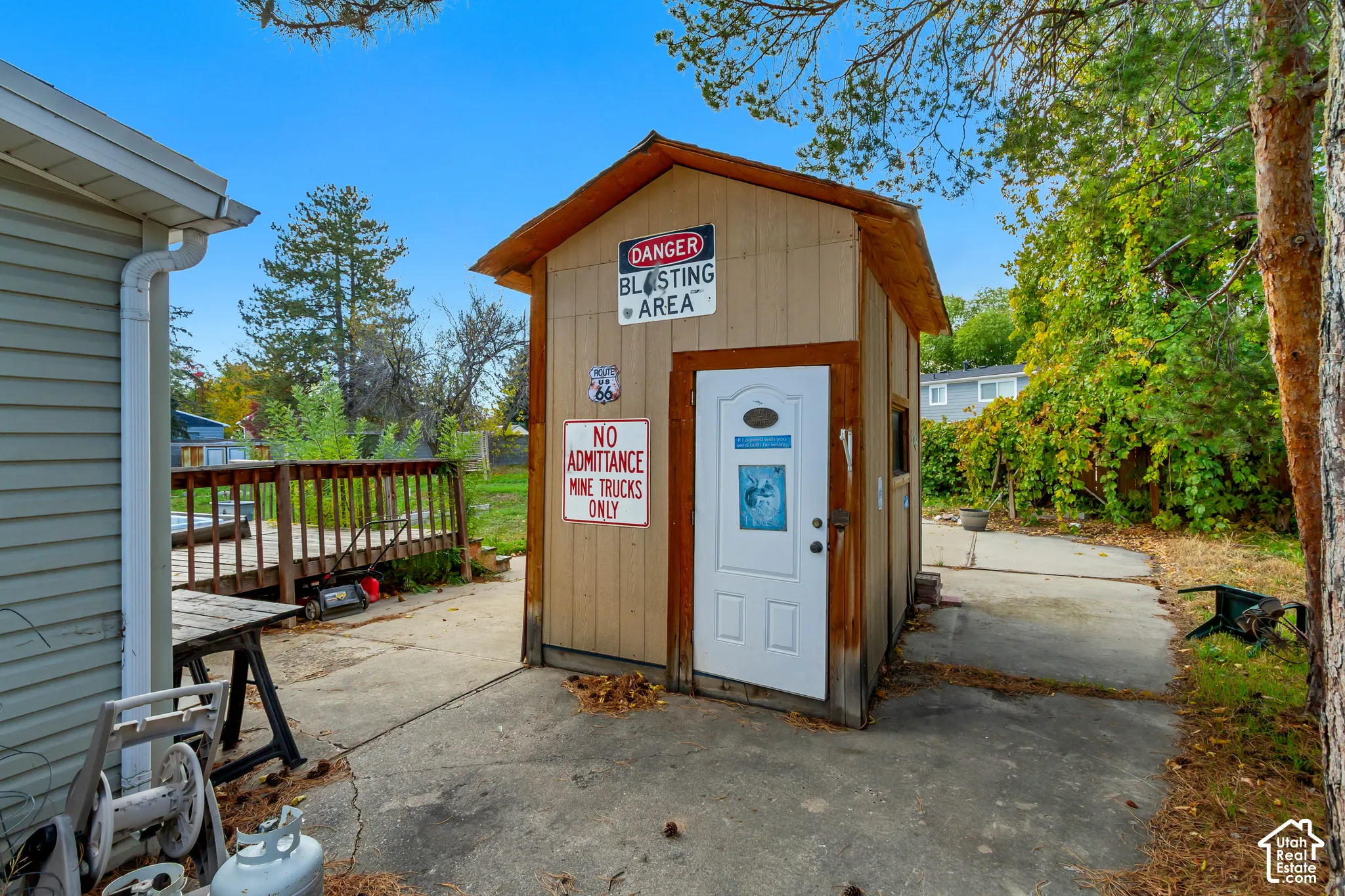 Exterior storage structure featuring wood-panel siding, a gabled roof, and a single entry door, set on a concrete pad with an elevated wood deck