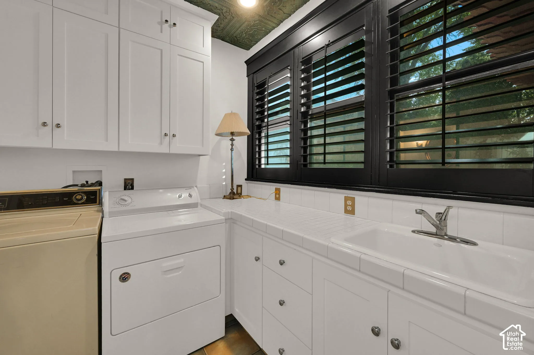 White shaker-style cabinetry, tiled countertop with an integrated utility sink, dark-finished window trim, and textured decorative ceiling