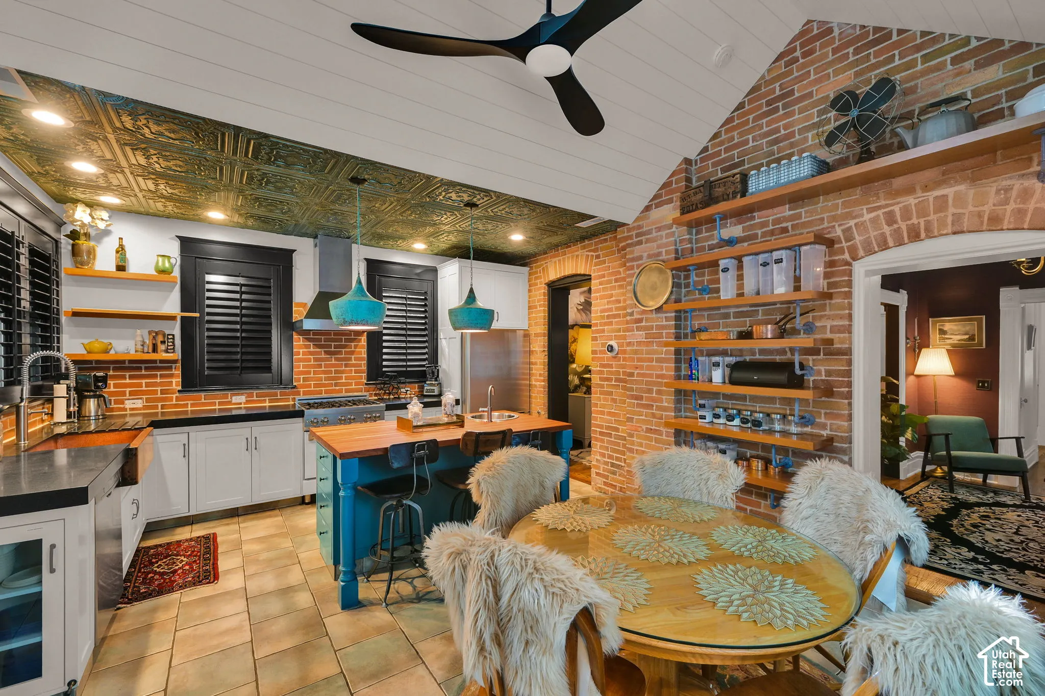 Vaulted shiplap ceiling with integrated ceiling fan, exposed red brick masonry walls, decorative metallic finish ceiling tiles, open wood shelving with industrial metal hardware, and center island featuring a wood countertop