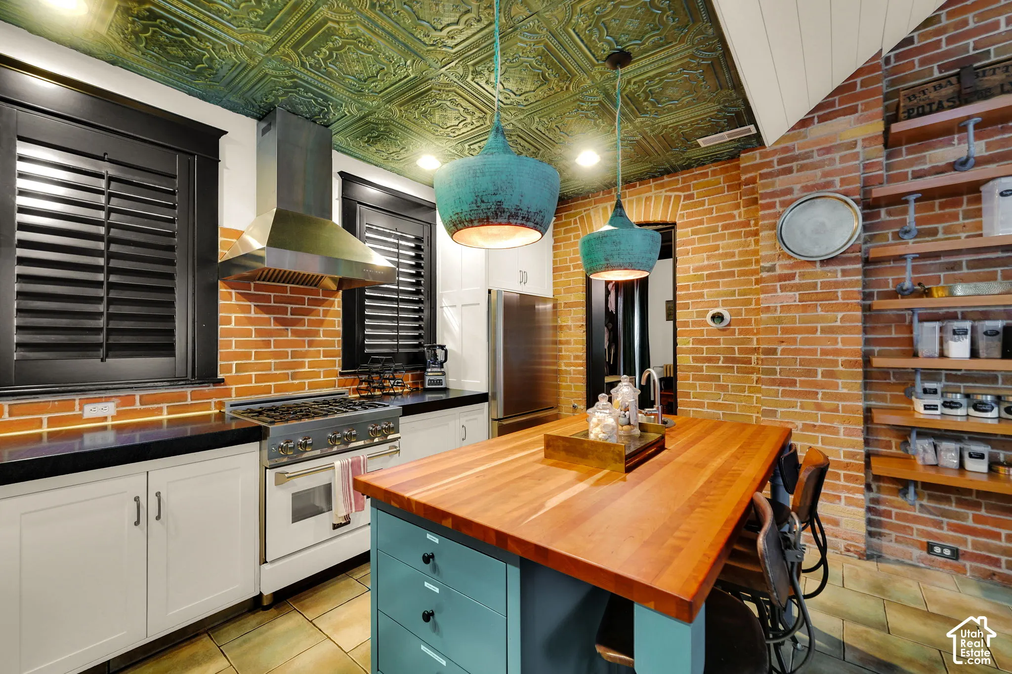 Kitchen featuring exposed brick walls, a custom stamped-metal ceiling, a butcher-block island, professional-style gas range with stainless steel hood, and industrial-style shelving