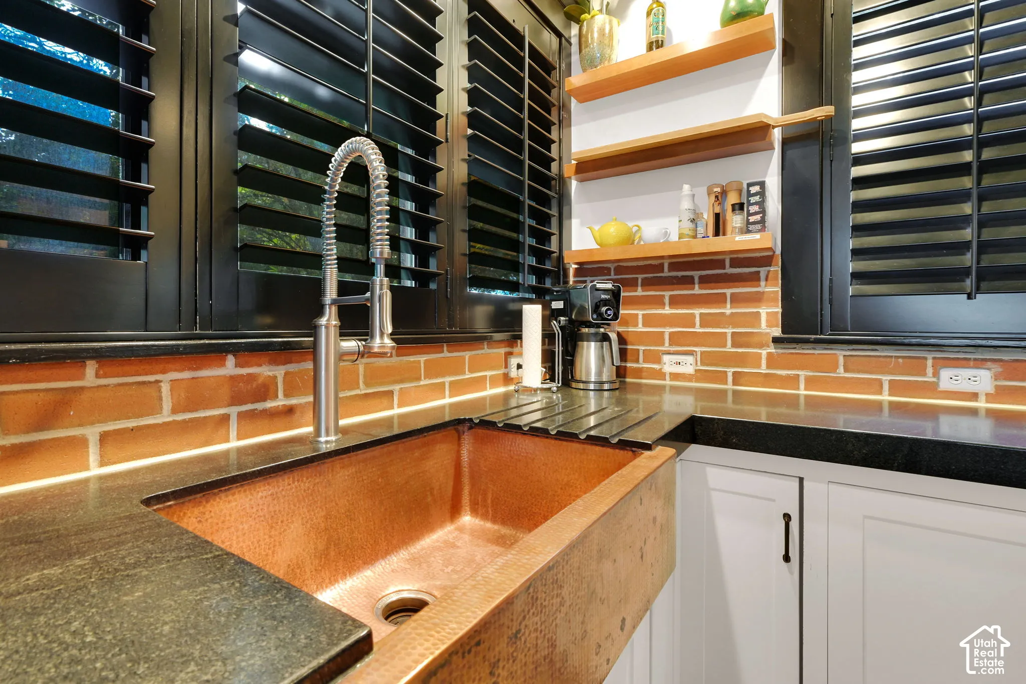 Copper farmhouse sink with a high-arch spring faucet, dark stone countertops, exposed brick backsplash, matte black window shutters, and wood-finish floating shelving