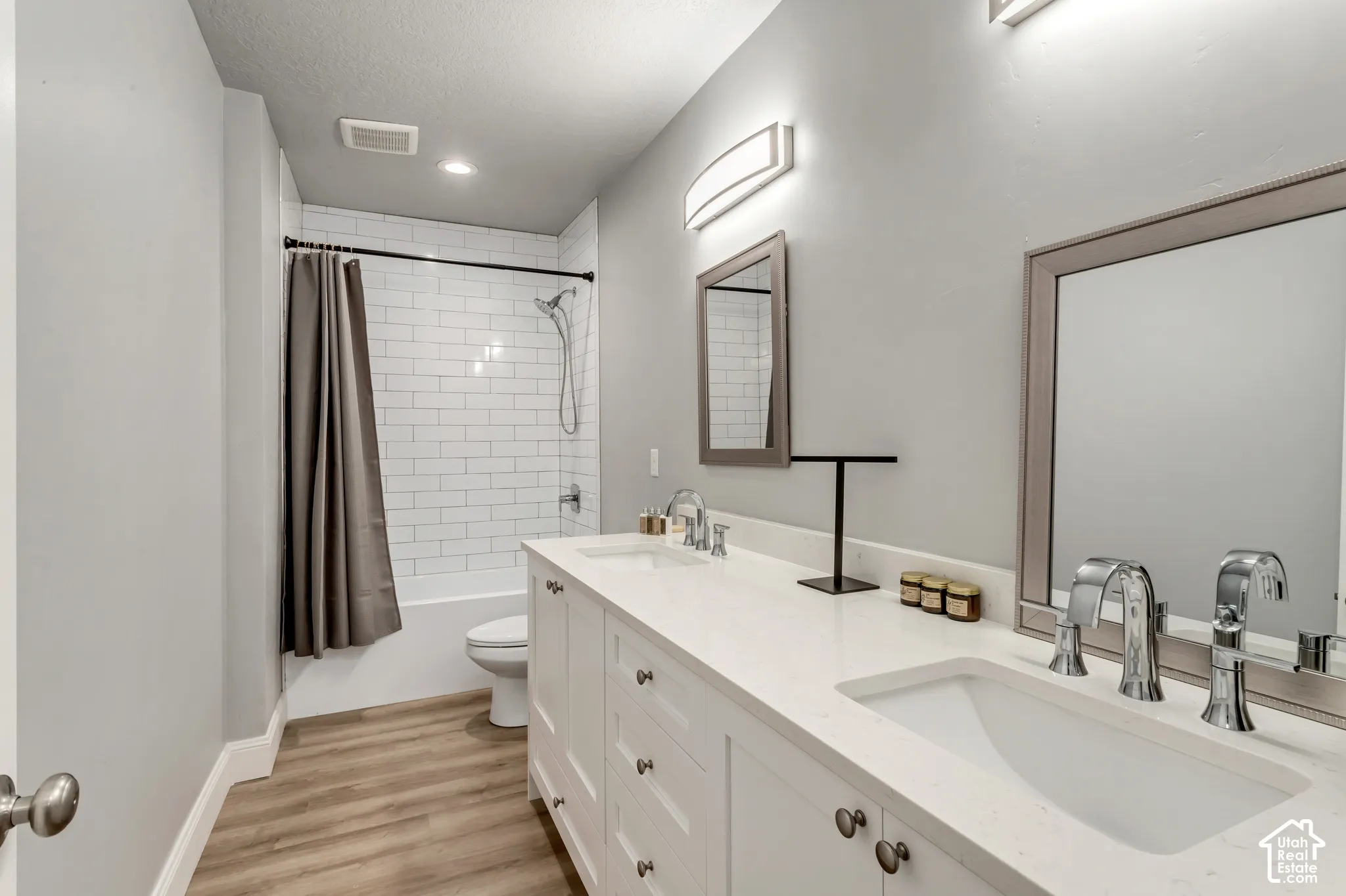 Dual-sink vanity with a quartz countertop, white shaker-style cabinetry, chrome fixtures, and a tub-shower combination with white subway tile surround, finished with wood-finish flooring