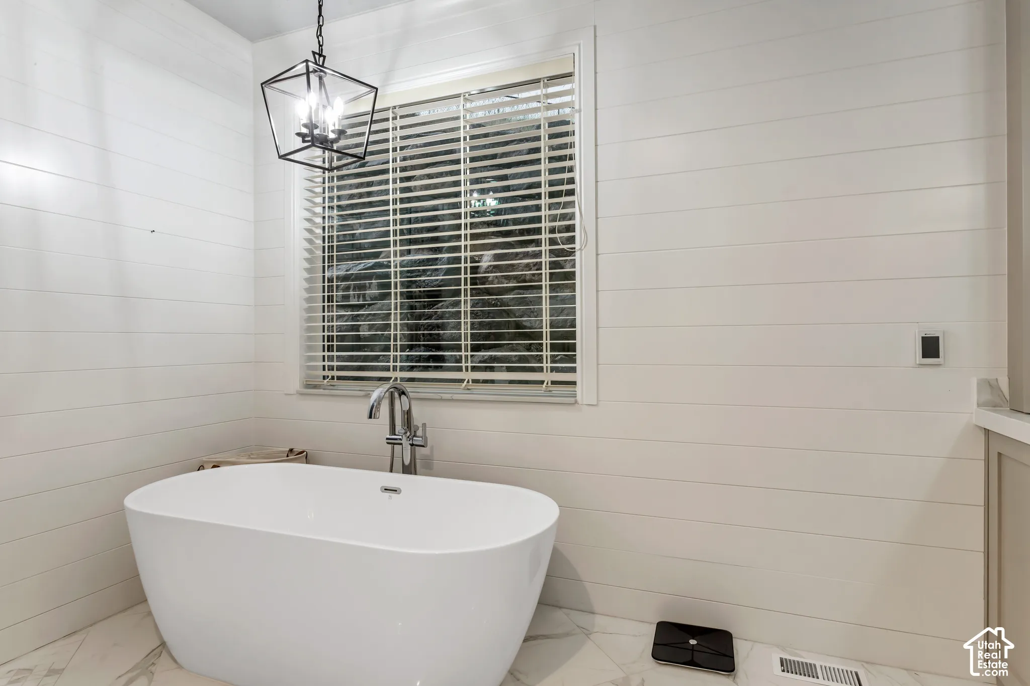 Freestanding white soaking tub with chrome floor-mounted faucet, horizontal white shiplap walls, patterned tile flooring, and geometric metal pendant lighting