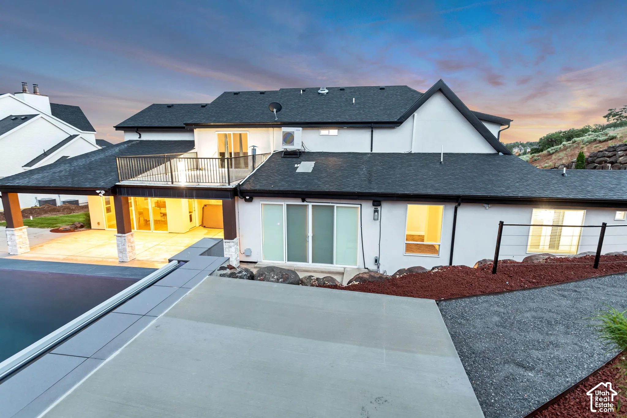 Rear exterior featuring a covered patio with stone-accented columns, second-story balcony with metal railings, sliding glass door access, and stone hardscaping