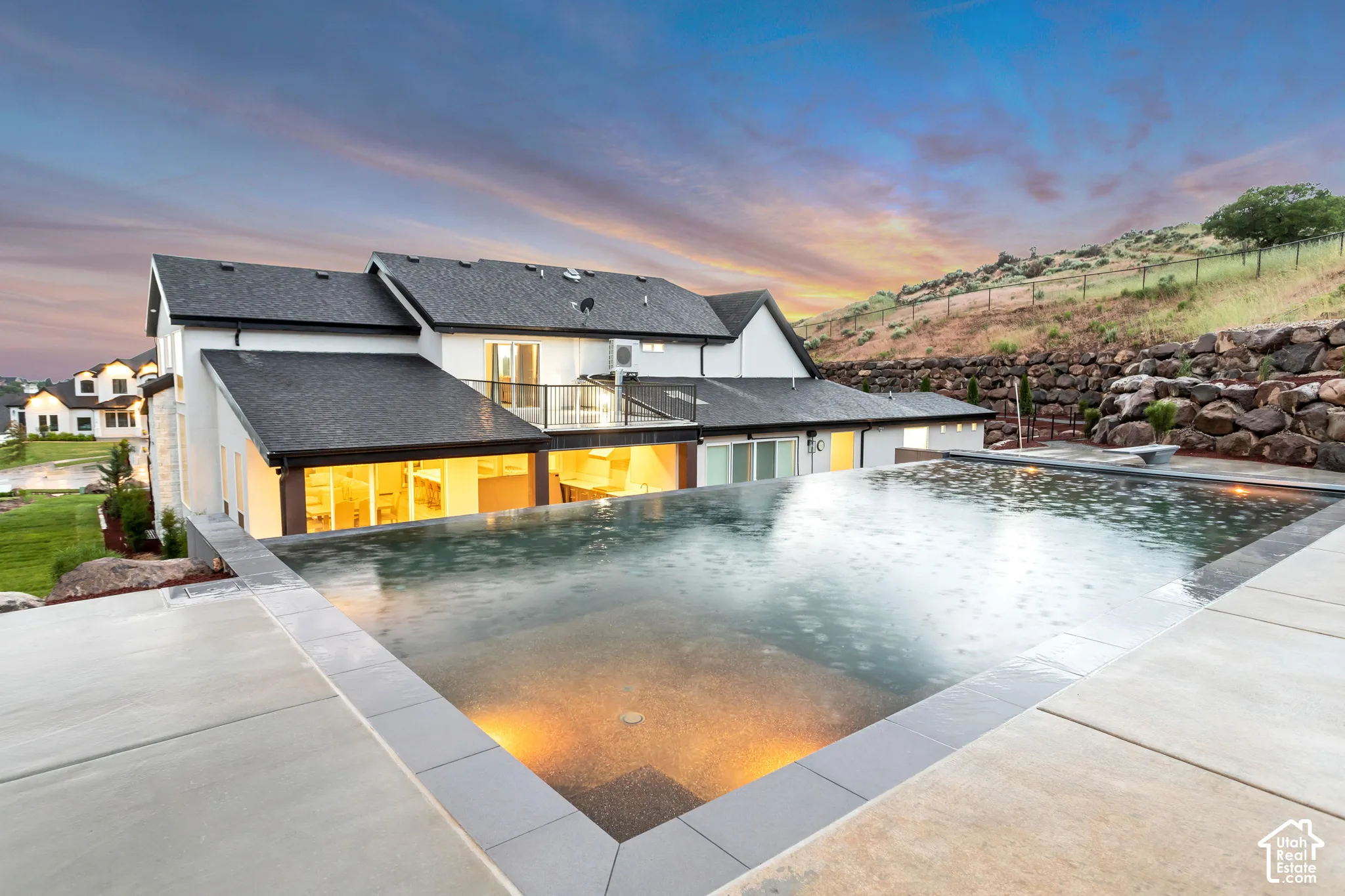 Infinity-edge pool with tiled surround, concrete patio, glass-railed second-story balcony, multi-gabled roofline, and stone retaining wall