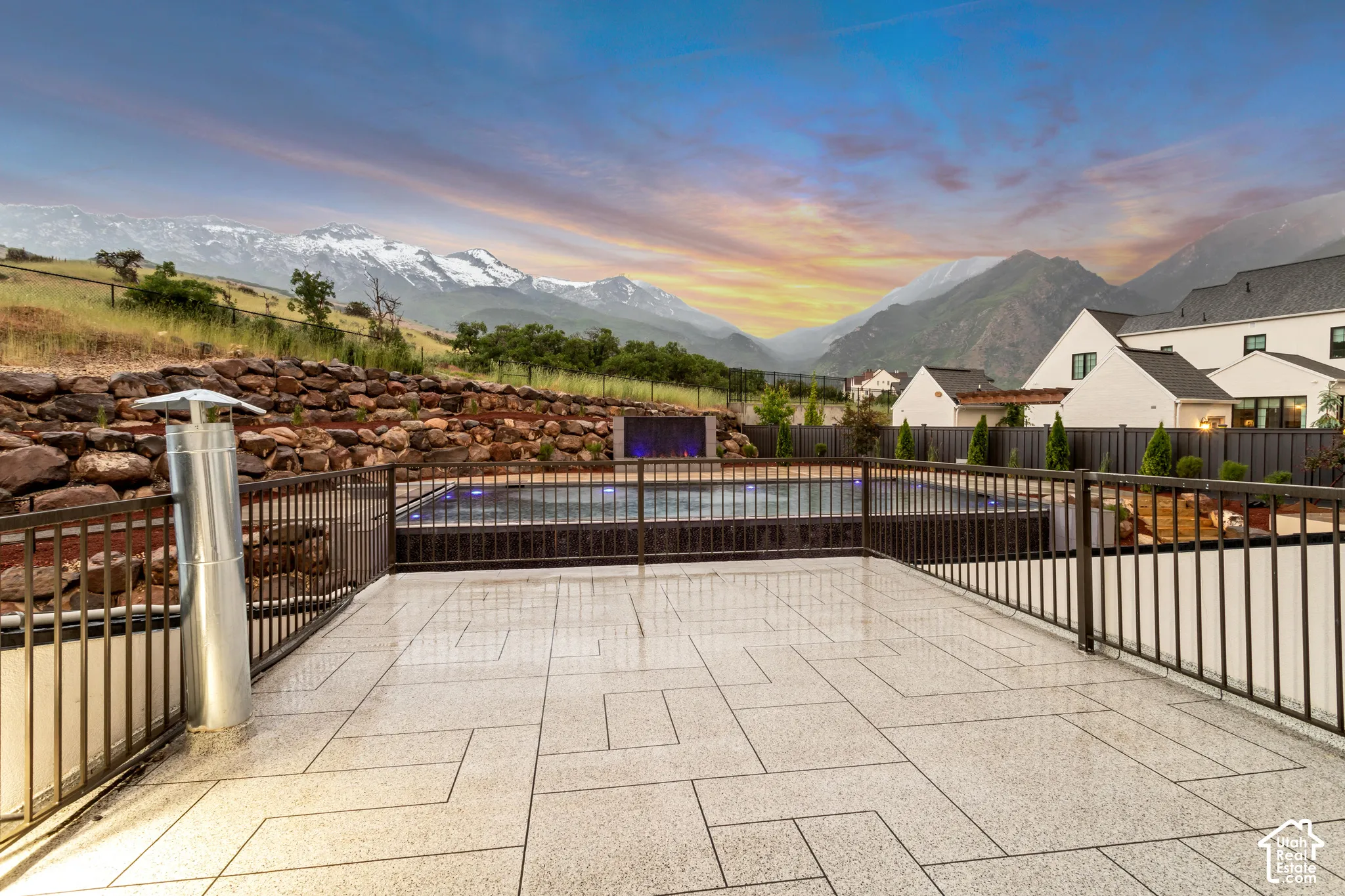 Stone-paver terrace featuring a dark metal railing, an infinity-edge swimming pool with a built-in water feature, and a stacked-stone retaining wall set against a mountain backdrop