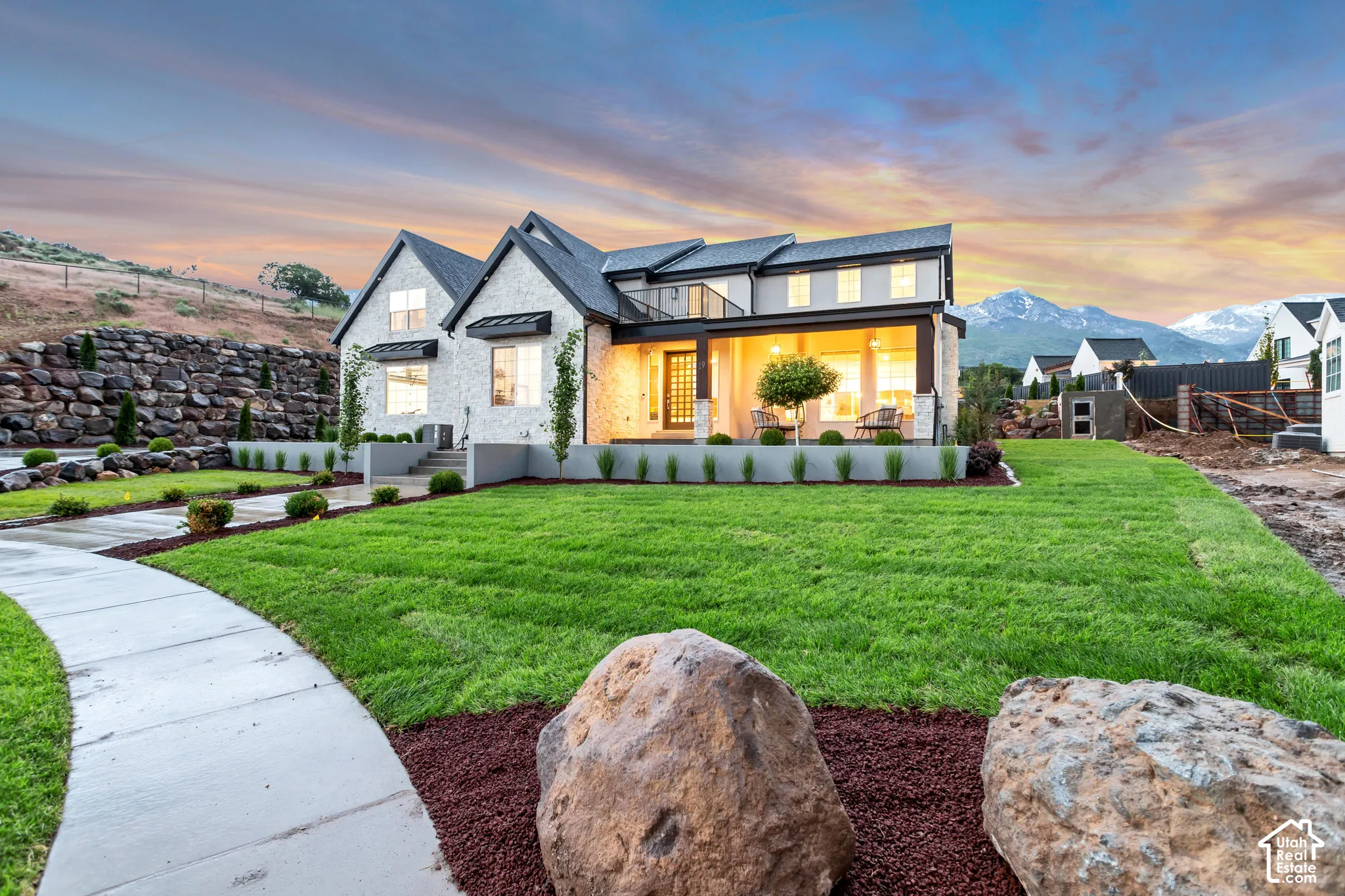 Two-story residence featuring a light-colored masonry facade, covered front porch with recessed lighting, black metal balcony railing, and landscaped grounds with a concrete walkway