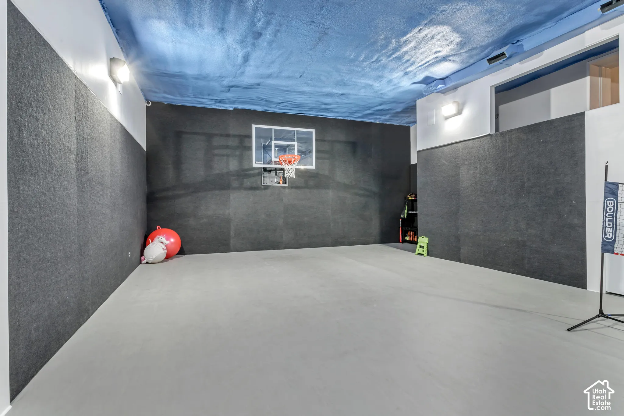 Indoor court featuring smooth flooring, dark textured wall paneling, a wall-mounted basketball hoop, and blue spray-foam ceiling insulation