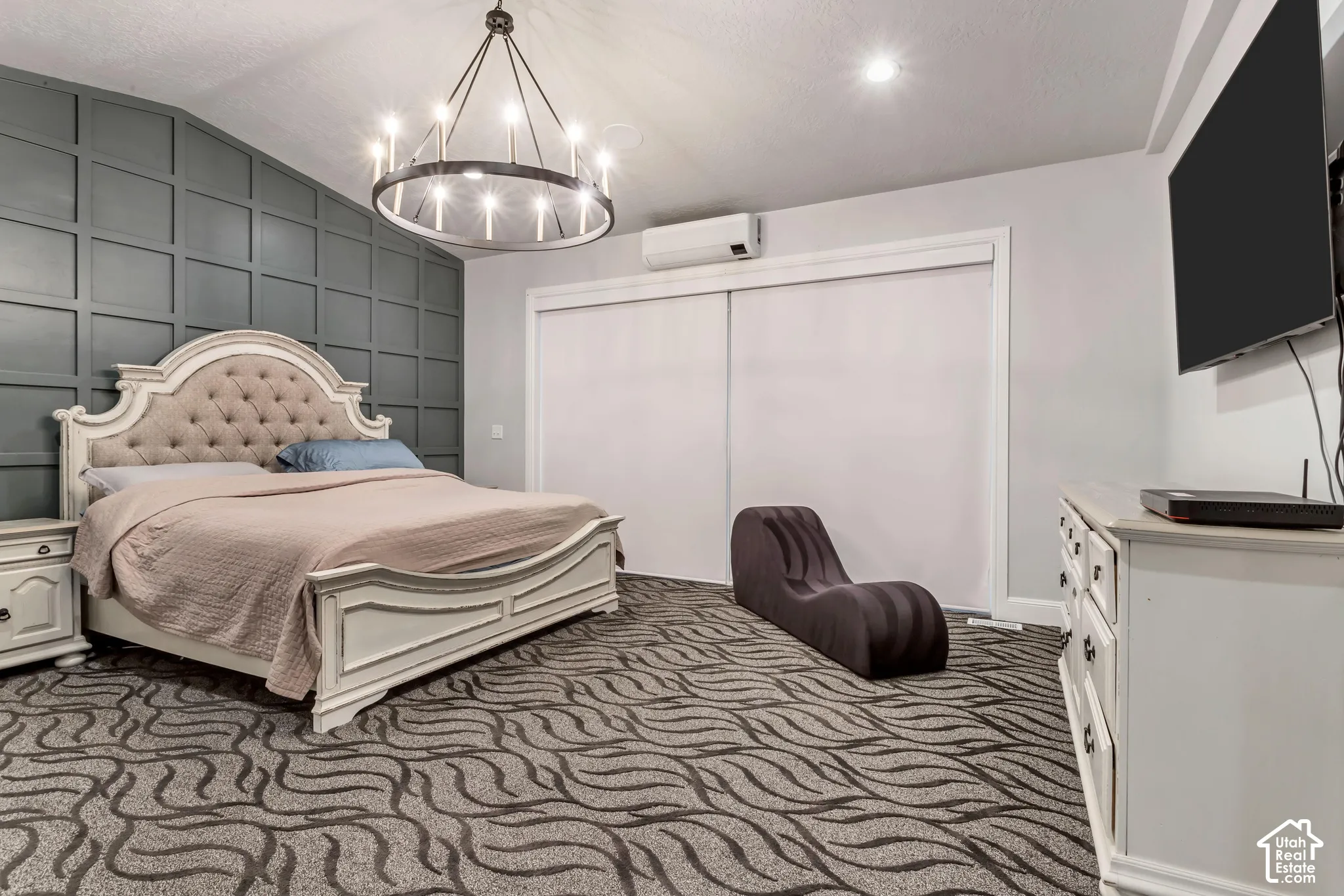 Room featuring a dark-toned square grid accent wall, a circular multi-arm chandelier, wall-mounted climate control unit, and sliding bypass closet doors