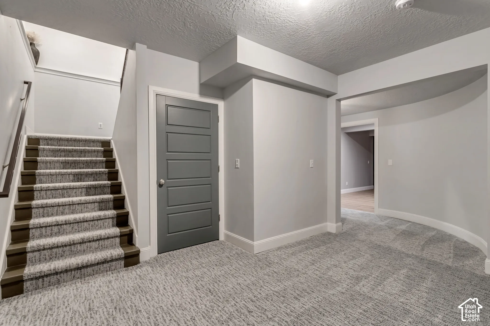 Interior space featuring grey-toned carpeting, staircase with wood-finish treads and metal handrail, five-panel door with metallic hardware, and curved wall architectural detail