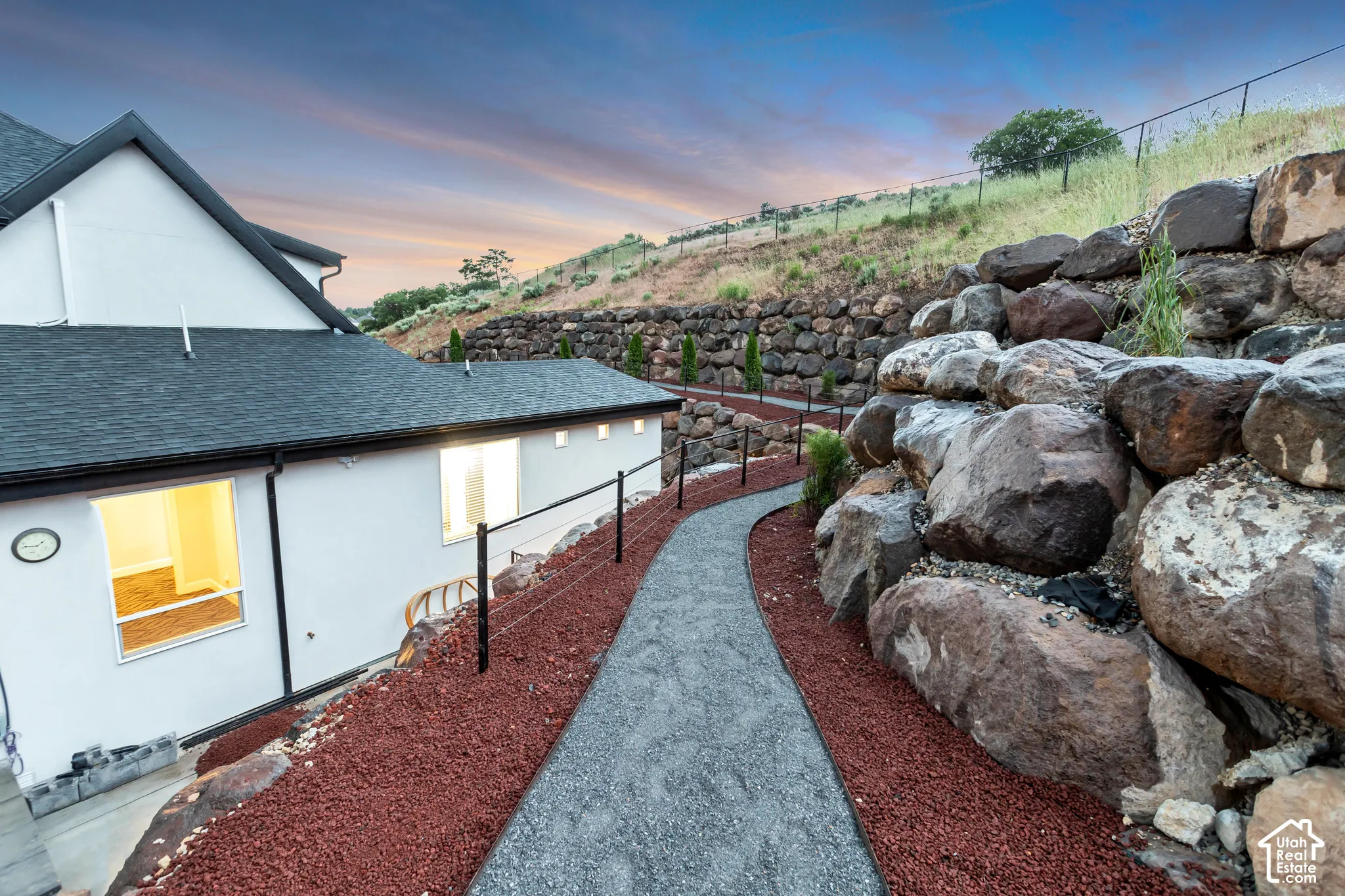 Exterior path featuring crushed stone gravel, tiered boulder retaining walls, and black metal post fencing against a hillside landscape