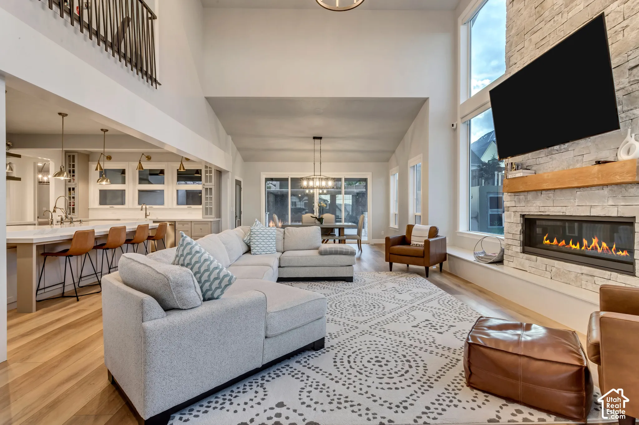Double-height ceilings, stone-clad fireplace with timber mantel, wood-finish flooring, recessed lighting, and kitchen island with pendant fixtures