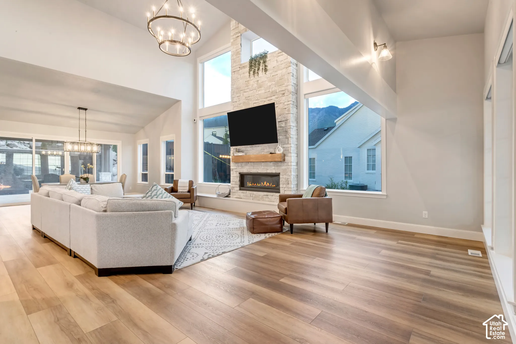 Vaulted ceilings, expansive stone-clad fireplace with timber mantel, wood-finish flooring, multi-pane windows, and modern chandelier lighting