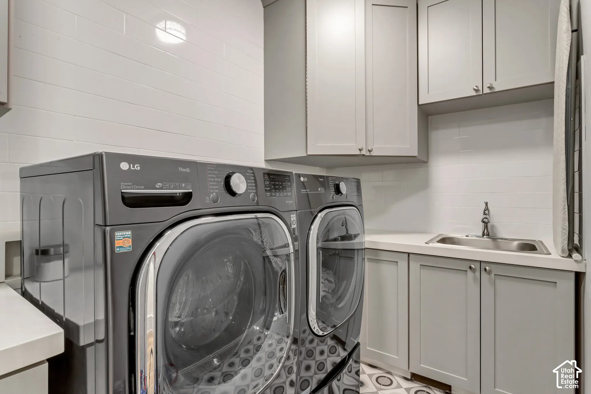 Laundry space featuring grey shaker-style cabinetry, a white countertop with an undermount sink, white tile backsplash, and patterned flooring