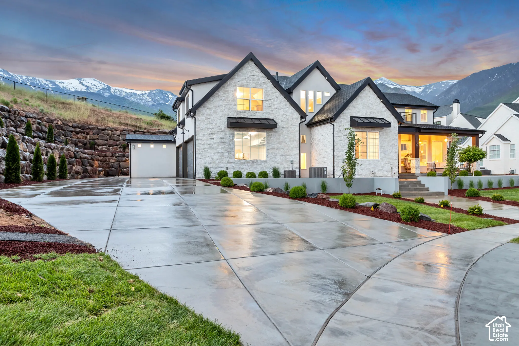 Multi-gabled exterior featuring white stone masonry, contrasting dark metal roofing accents, an expansive concrete driveway, and a mountain backdrop