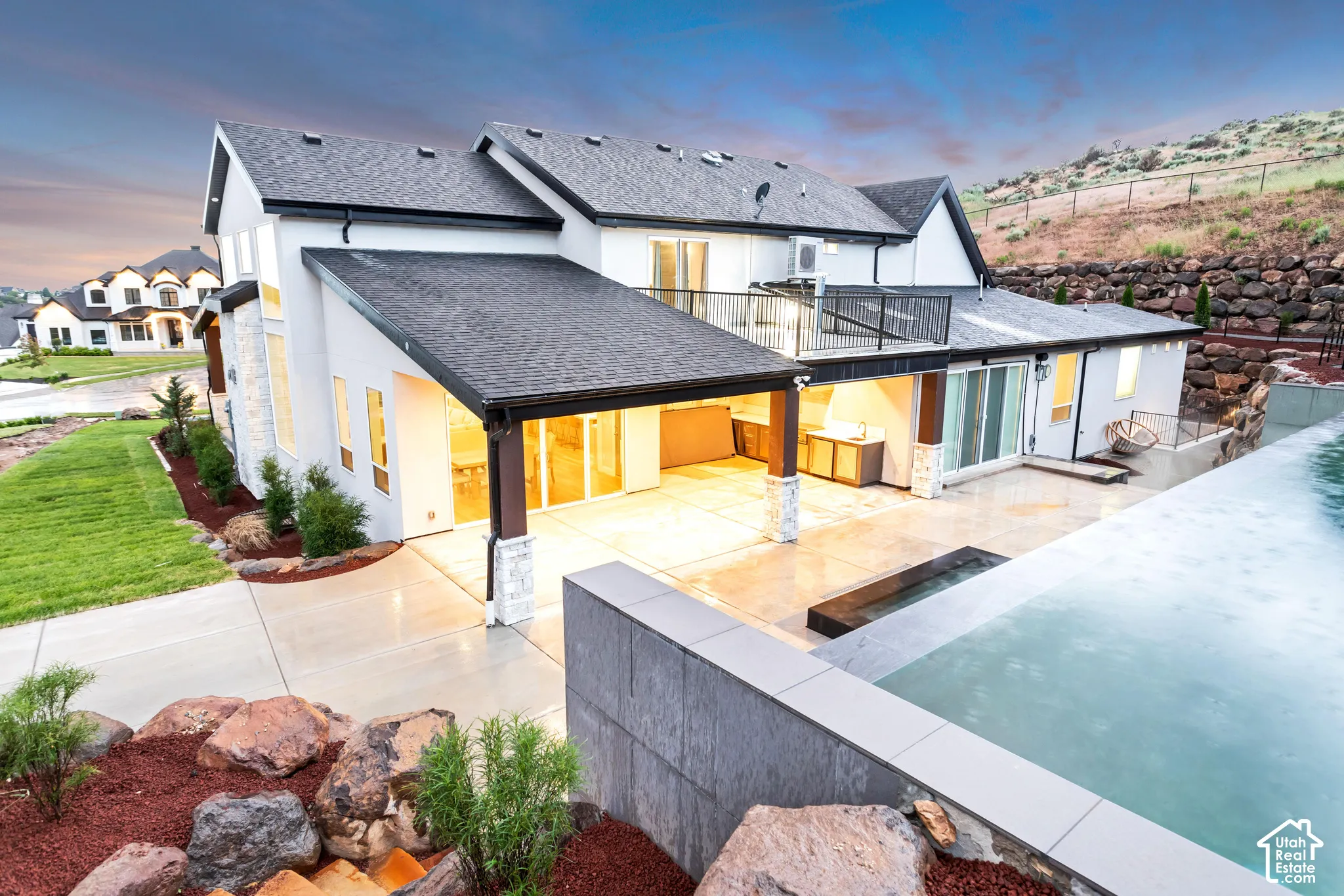 Multi-level exterior featuring a covered concrete patio, second-story balcony with metal railing, infinity-edge swimming pool, and stone-accented columns