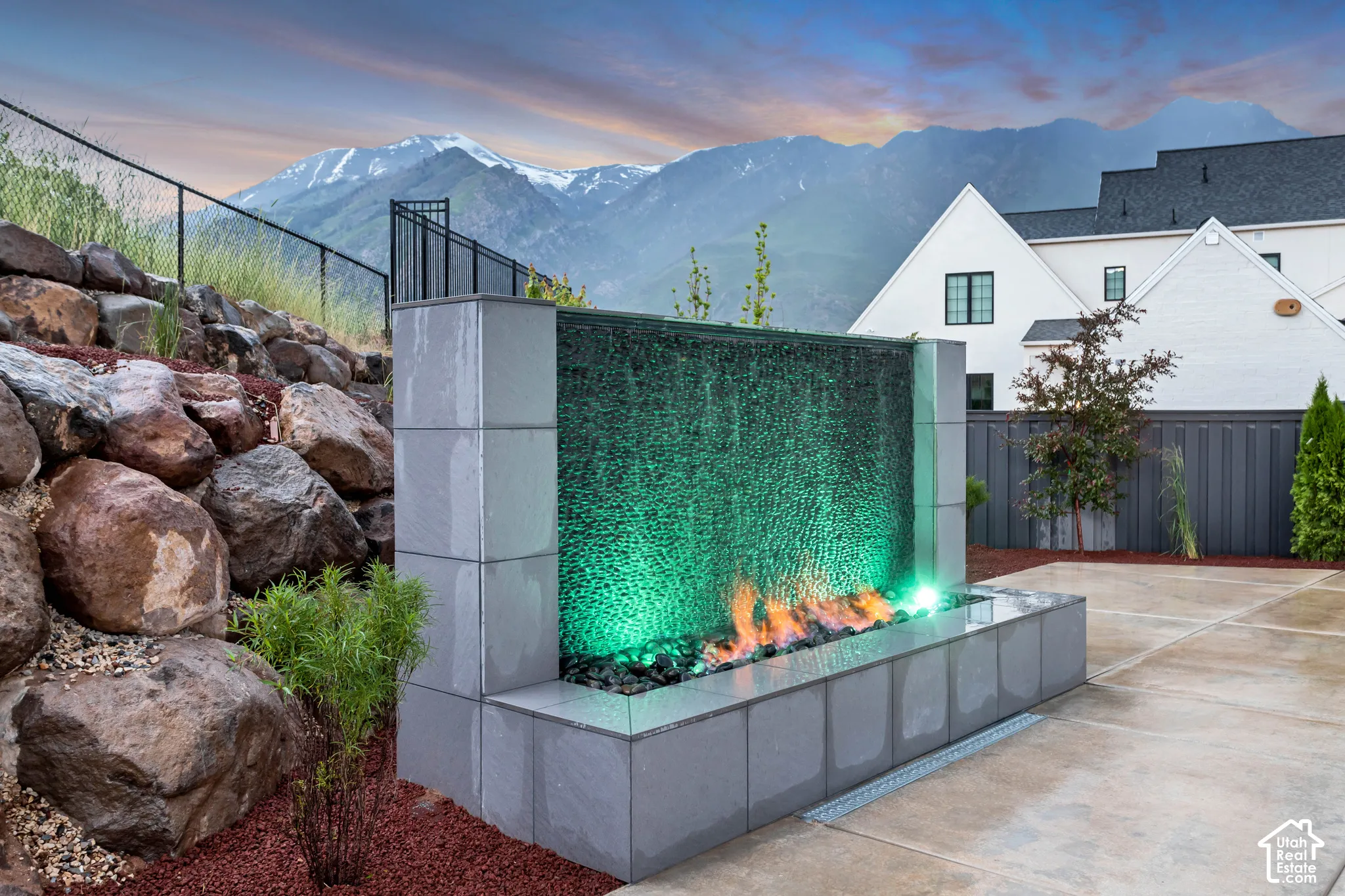 Custom stone water feature with integrated fire element, concrete patio, mountain backdrop, and tiered rock landscaping