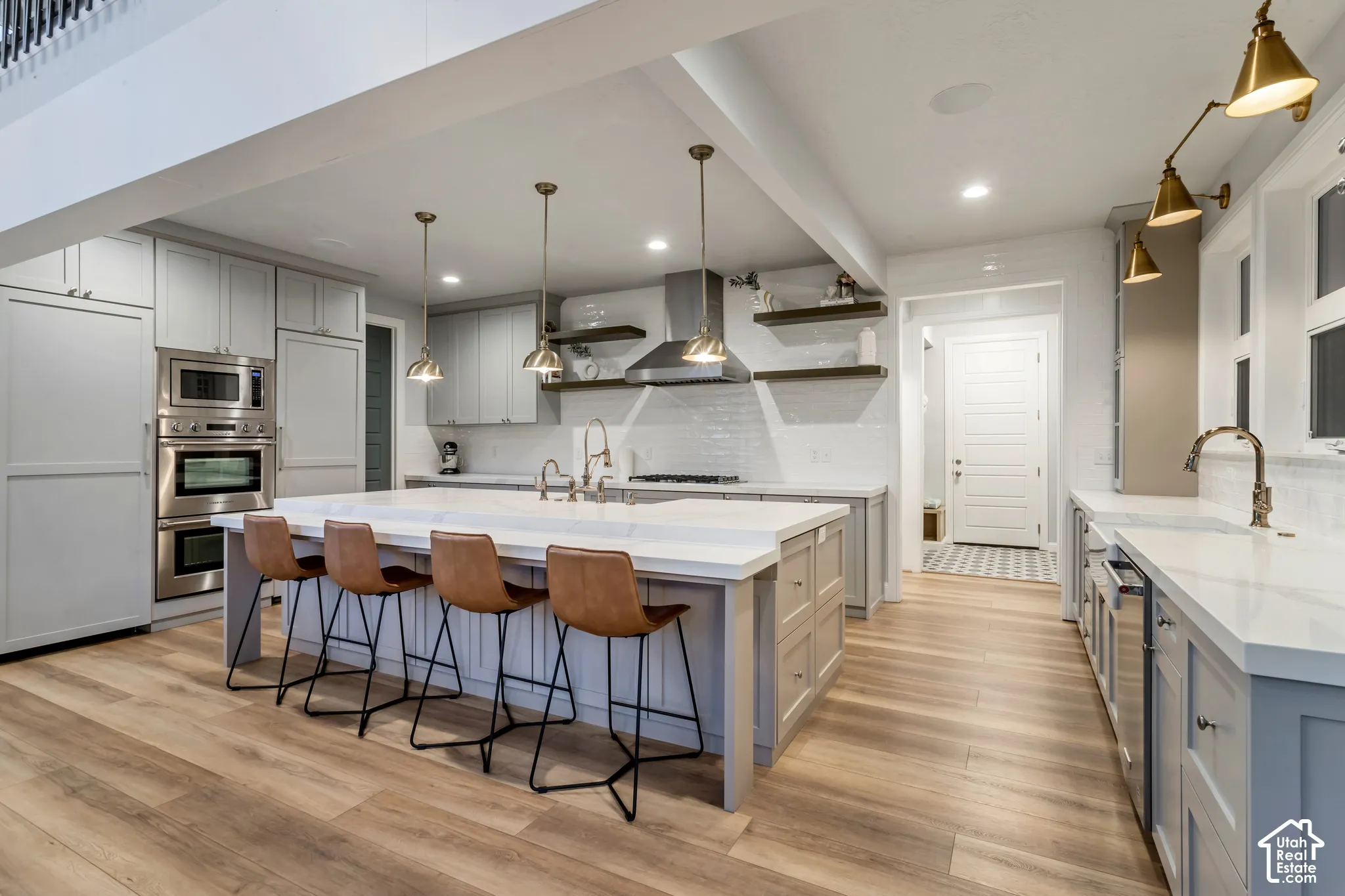 Kitchen featuring an oversized island with a waterfall edge, stainless steel double wall ovens, floating shelving, pendant lighting, and wood-finish flooring