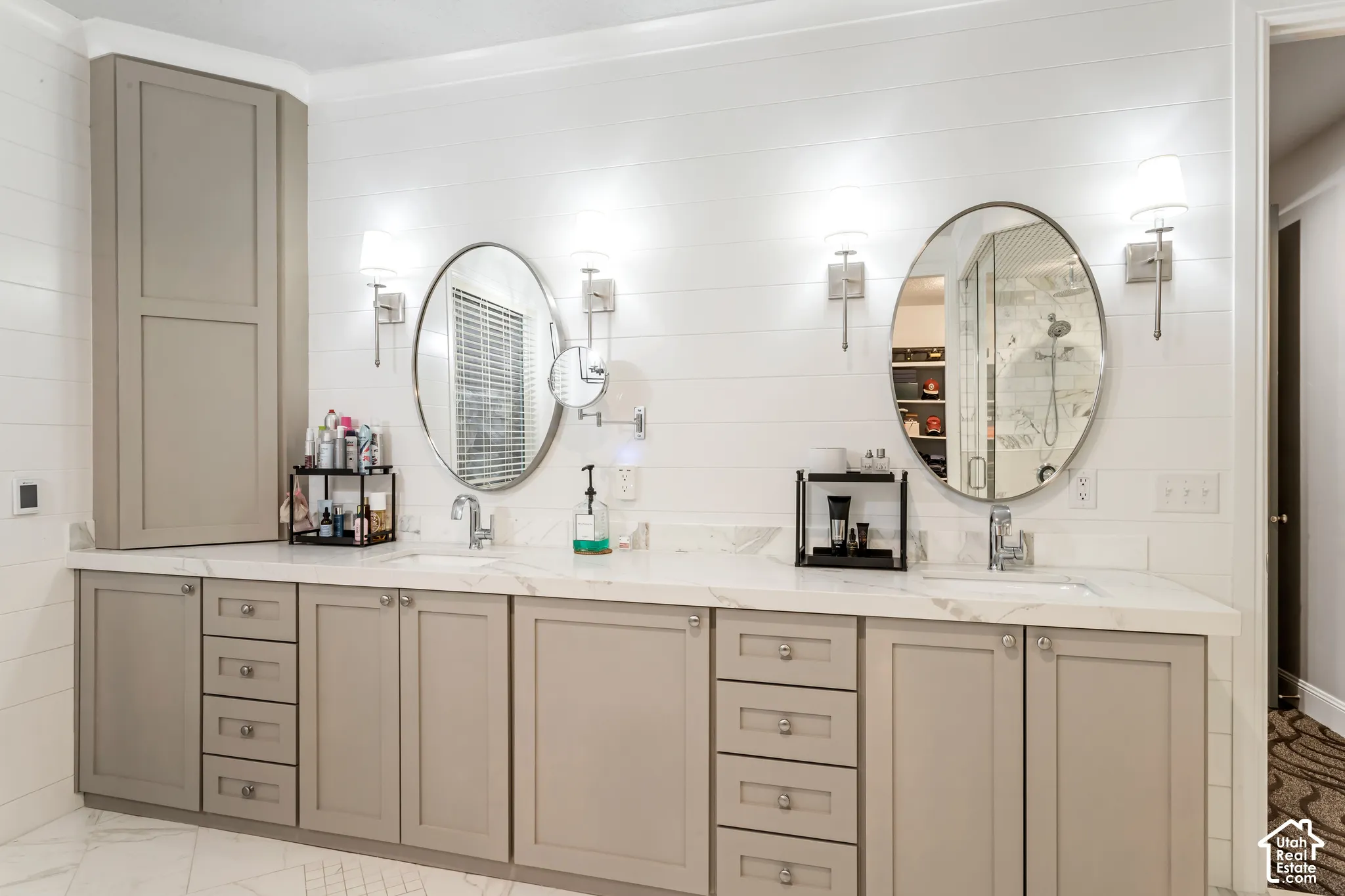 Dual-sink vanity featuring neutral-toned cabinetry, white stone-finish countertops, wall-mounted light fixtures, oval mirrors, and shiplap wall paneling