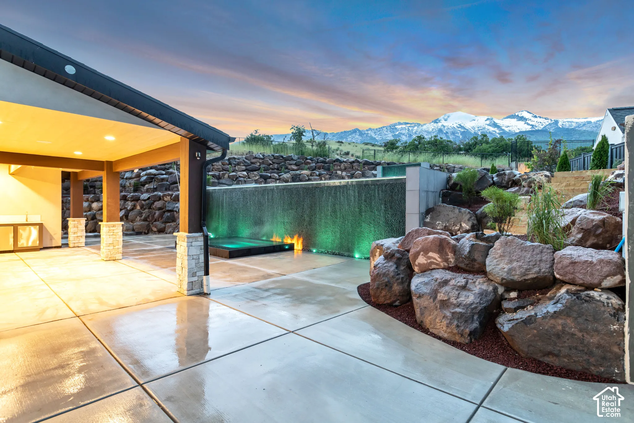 Covered patio featuring stone-clad pillars, a built-in water feature, integrated in-ground spa, and tiered natural rock landscaping, set against a backdrop of snow-capped mountains