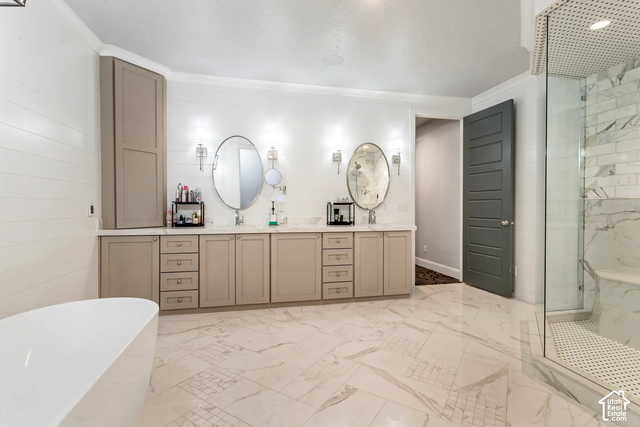 Bathroom featuring dual-sink vanity with beige cabinetry, oval wall-mounted mirrors, freestanding soaking tub, stone-patterned tile flooring, and glass-enclosed shower with tiled surround