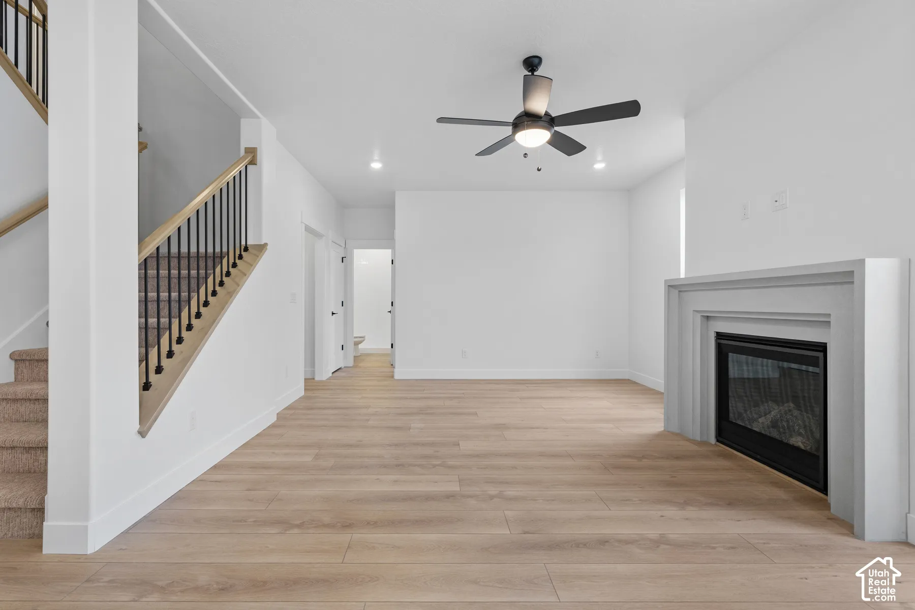 Open-concept interior featuring wood-finish flooring, a fireplace with a minimalist surround, a modern ceiling fan with integrated lighting, and a staircase with a light wood handrail and dark metal balusters