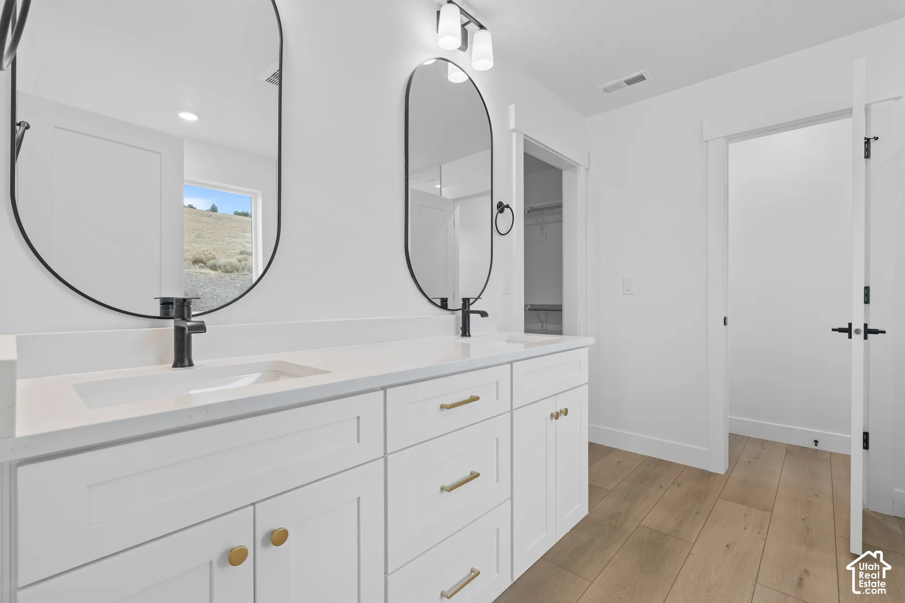 Dual-sink vanity with a solid-surface countertop, arched black-framed mirrors, matte black fixtures, and shaker-style cabinetry with gold-tone hardware, complemented by wood-finish flooring