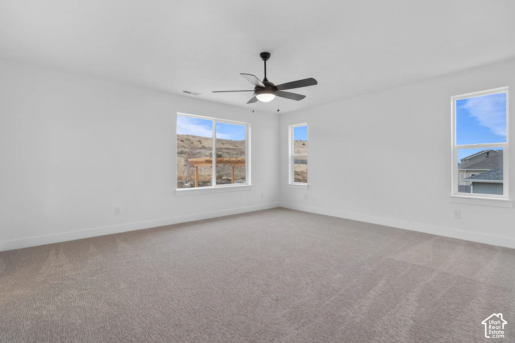 Neutral-toned wall-to-wall carpeting, three multi-pane windows, and a matte black ceiling fan with integrated lighting