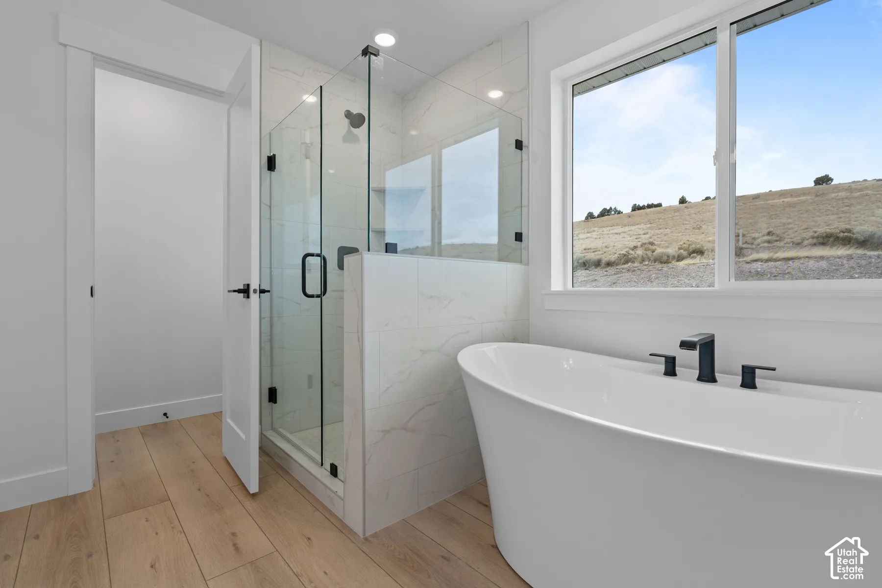 Freestanding soaking tub with matte black faucet, glass-enclosed shower featuring stone-patterned tile surround, recessed lighting, and wood-finish flooring