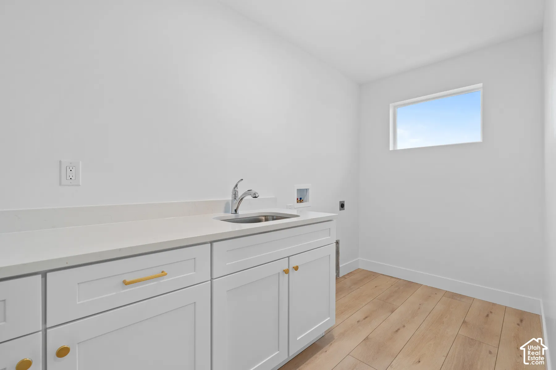 Laundry area featuring white shaker cabinetry, gold-tone hardware, a quartz countertop with an integrated basin, a polished chrome pull-down faucet, and light wood-finish flooring