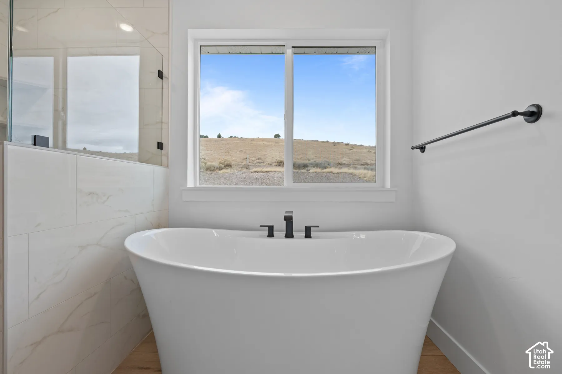 Freestanding soaking tub featuring matte black deck-mounted hardware, polished stone-finish wall tile, sliding window, and matte black towel bar