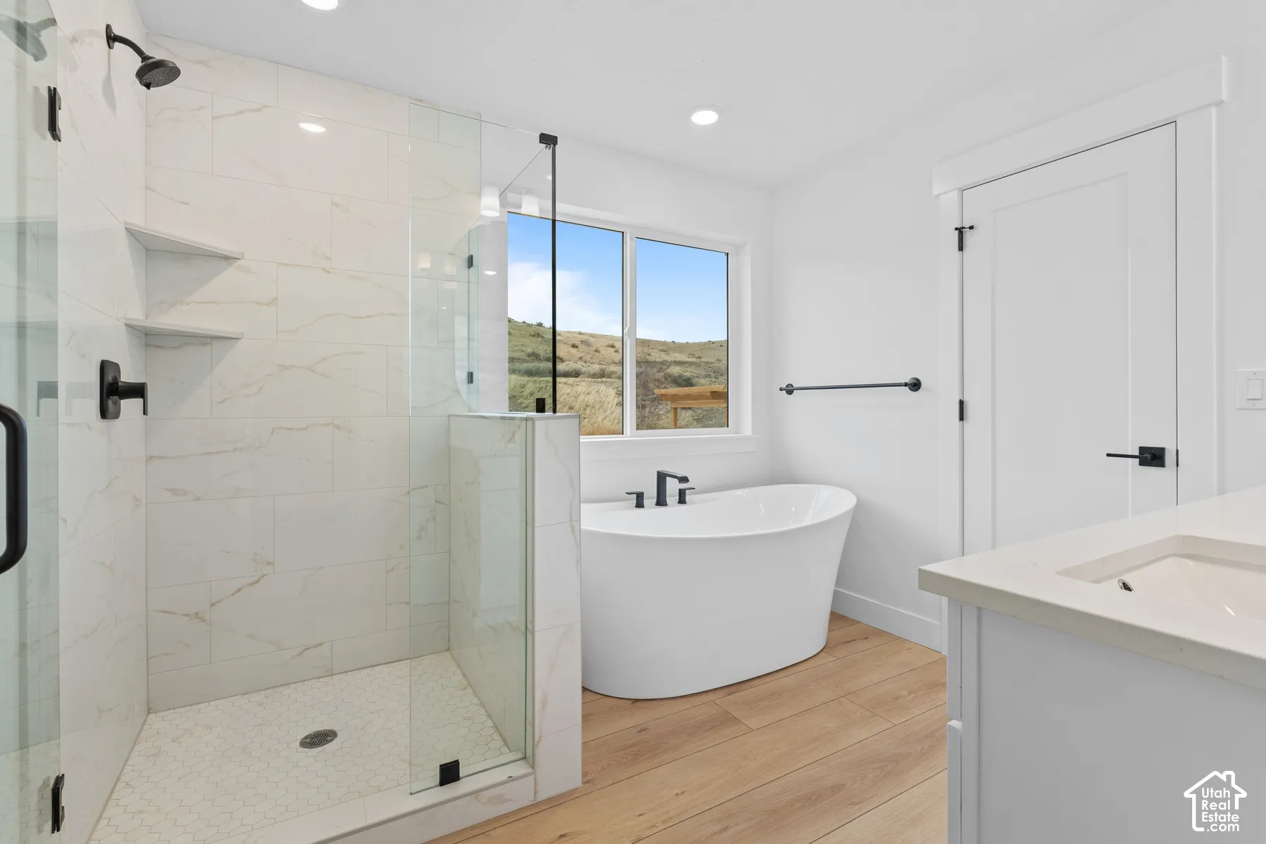 Walk-in shower with floor-to-ceiling tile, freestanding soaking tub, wood-finish flooring, matte black fixtures, and solid-surface vanity countertop