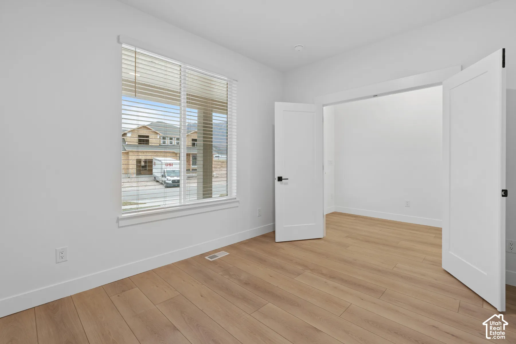 Room interior featuring light-tone wood-finish flooring, white walls, and a window with horizontal blinds