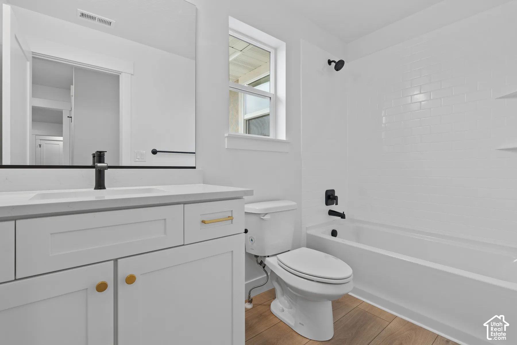 Bathroom featuring a white vanity with gold-tone hardware, wood-finish flooring, subway-tile tub surround, and matte black fixtures
