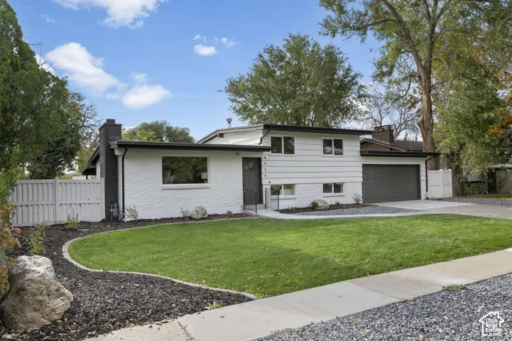 Split-level exterior featuring white painted brick and horizontal siding, a dark-toned attached garage, a chimney with dark masonry, and a landscaped front lawn with mulch borders
