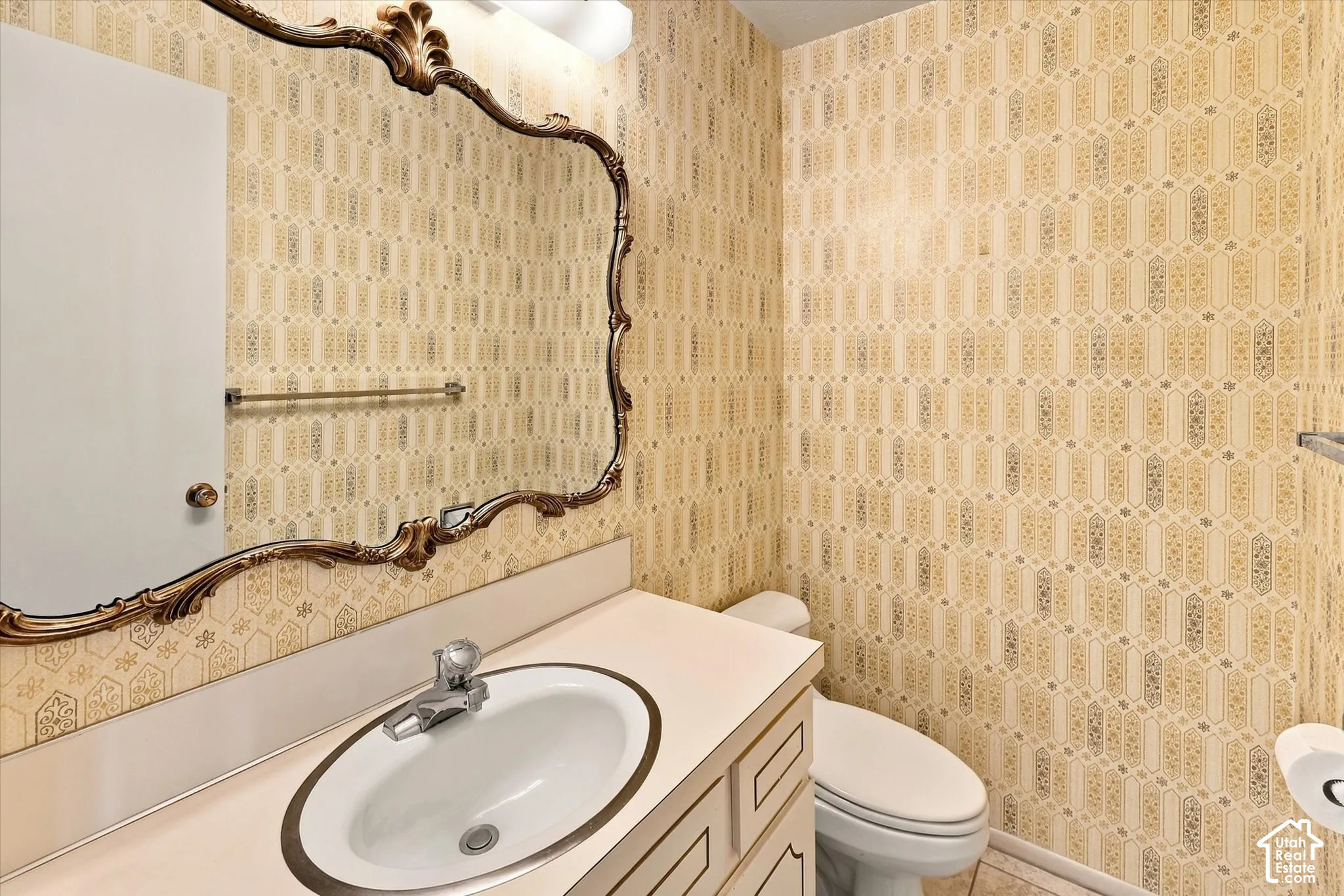 Bathroom featuring patterned wallpaper, a vanity with an oval inset sink, an ornate wall mirror, a toilet, and an overhead light fixture