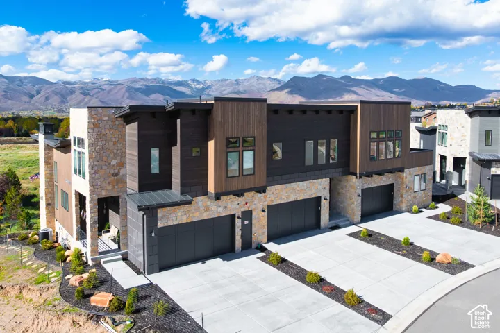 Contemporary multi-unit exterior featuring stacked stone masonry, dark metal-finish accents, horizontal wood-finish siding, attached multi-car garages, and mountain terrain backdrop
