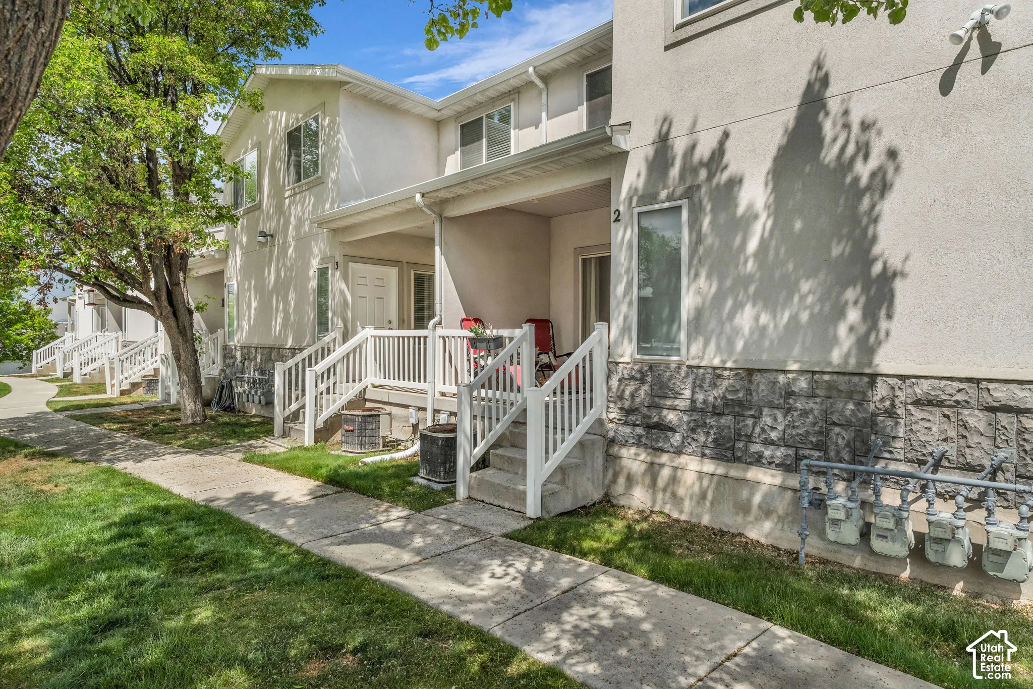 Multi-level exterior featuring stucco siding, stone veneer lower-level cladding, covered porches with white railing, and concrete walkways