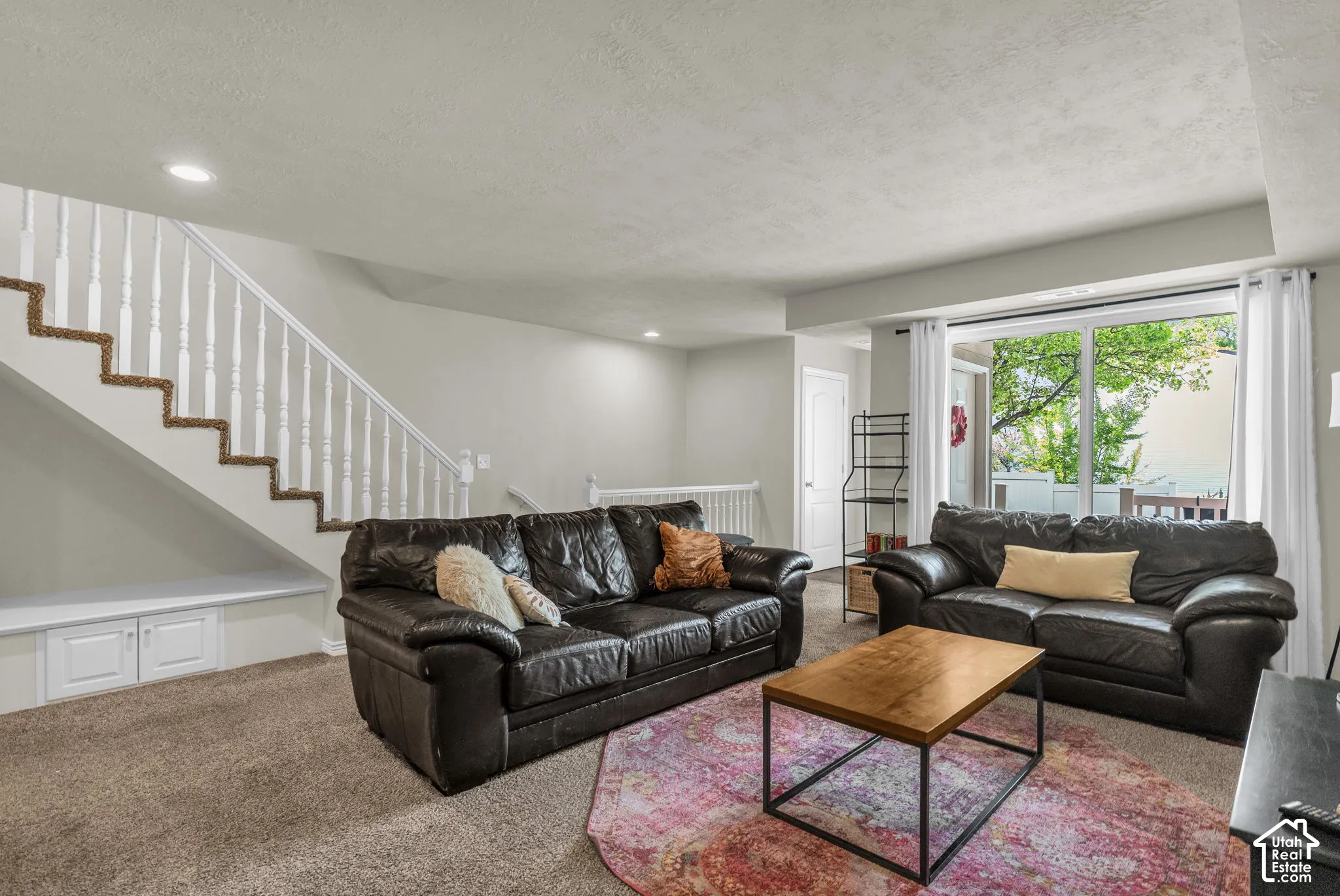 Open floor area featuring neutral wall tones, carpeted flooring, recessed lighting, and a staircase with white balusters and built-in under-stair storage cabinetry