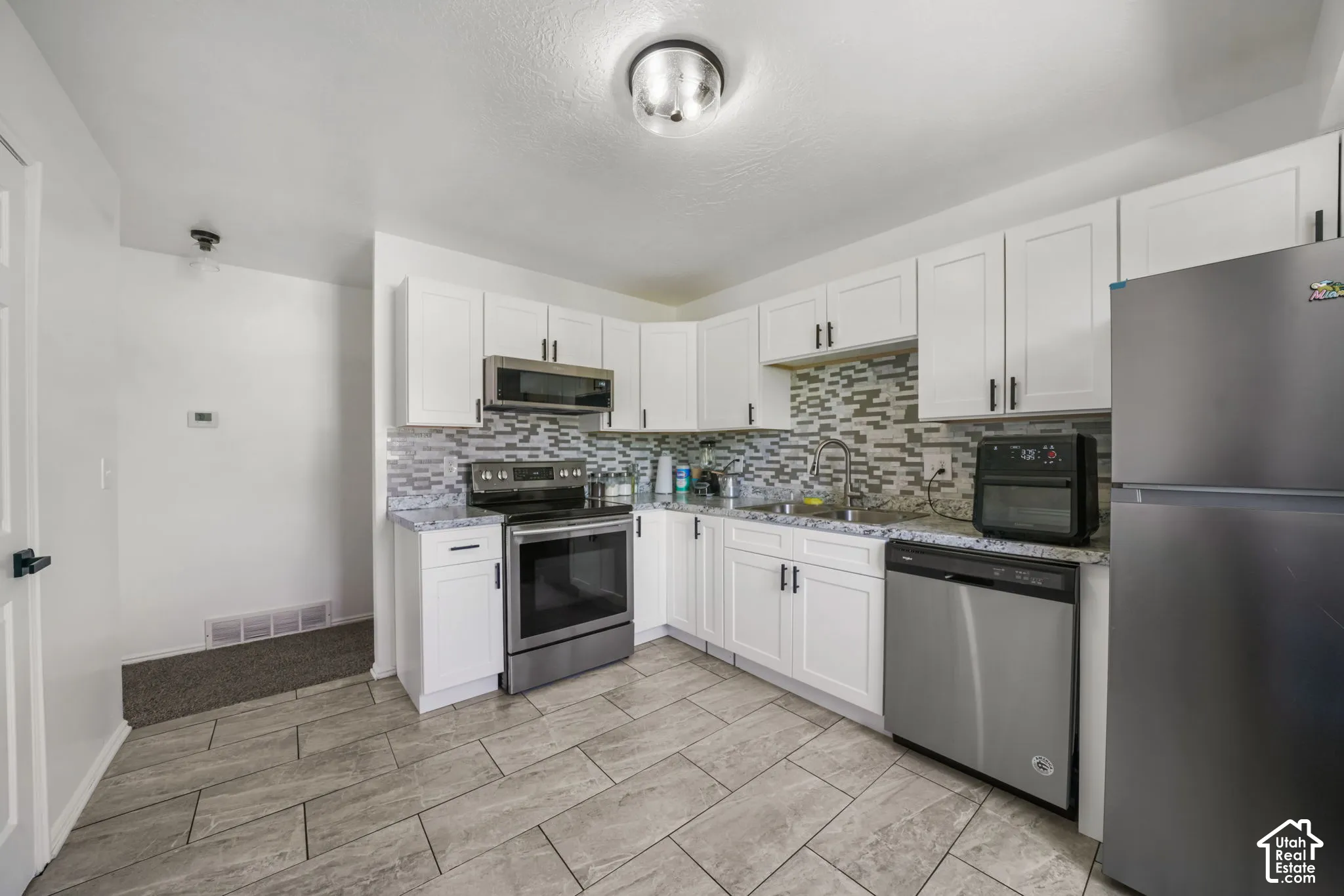 Kitchen featuring white shaker cabinetry, stone-patterned countertops, a multi-tone tile backsplash, stainless steel appliances, and large-format tile flooring