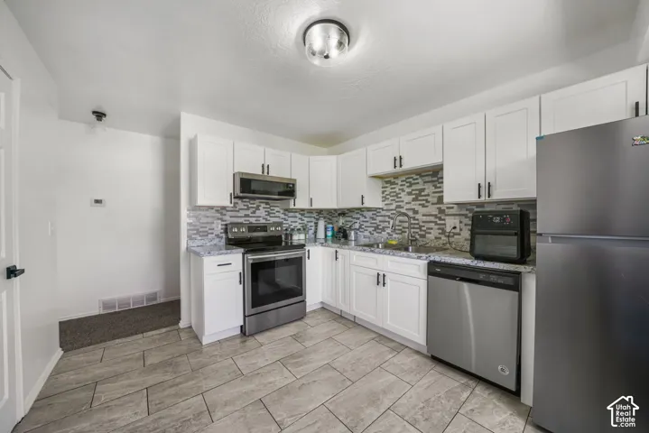 Kitchen featuring white shaker cabinetry, stone-patterned countertops, a multi-tone tile backsplash, stainless steel appliances, and large-format tile flooring