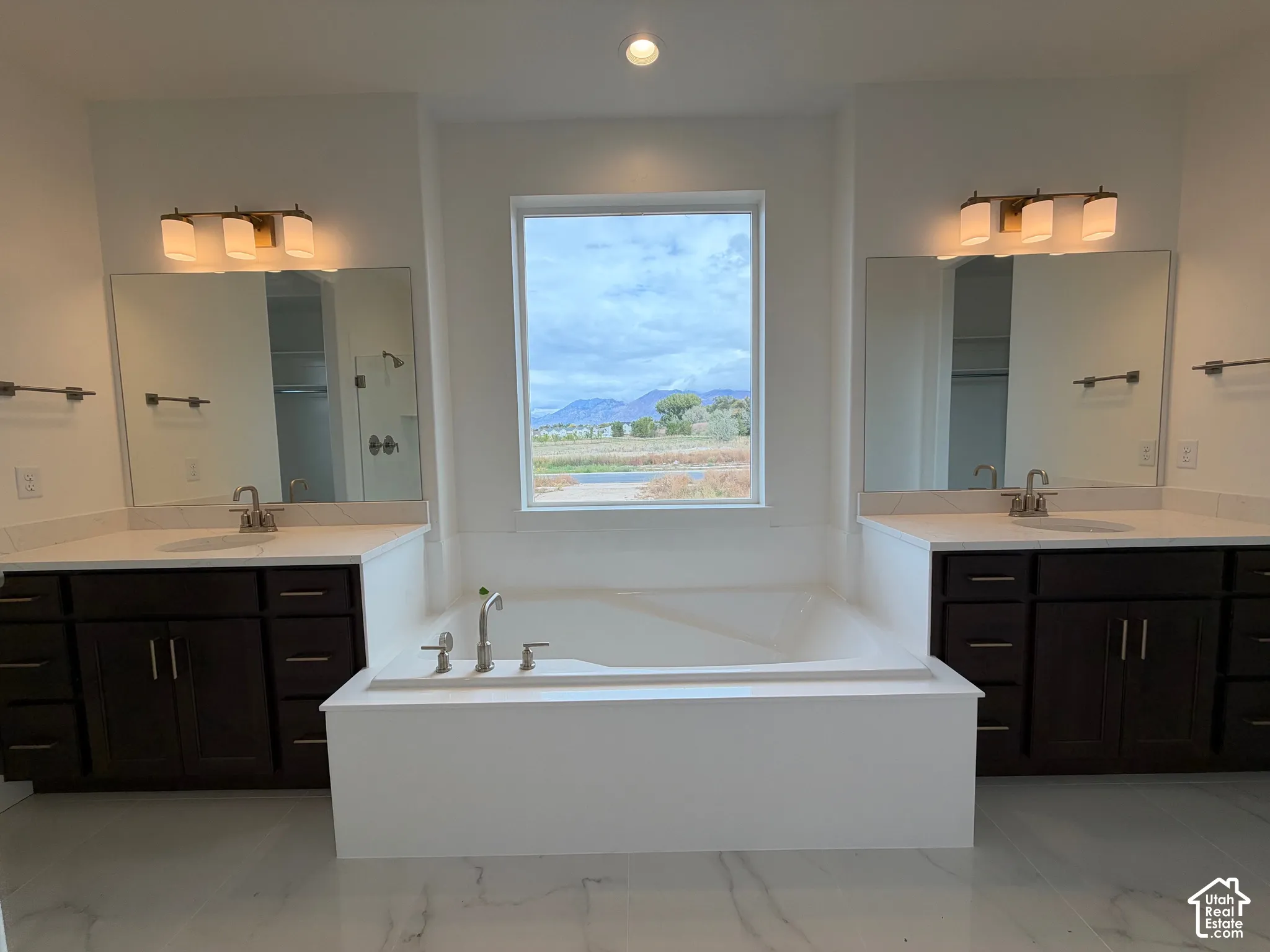 Dual vanities featuring dark cabinetry, solid-surface countertops, and integrated mirrors, centered by a deep soaking tub and a mountain backdrop