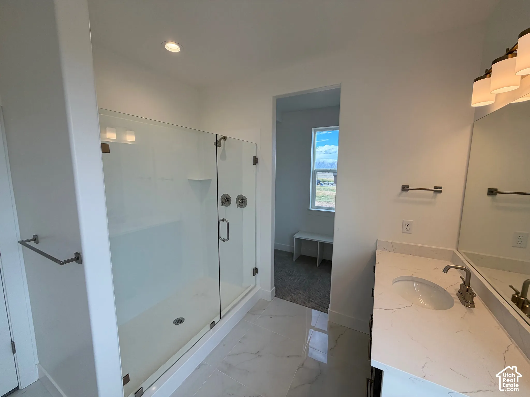 Bathroom featuring a walk-in shower with glass enclosure, light-toned stone-finish countertop, dual-basin vanity, and large wall-mounted mirror with integrated lighting
