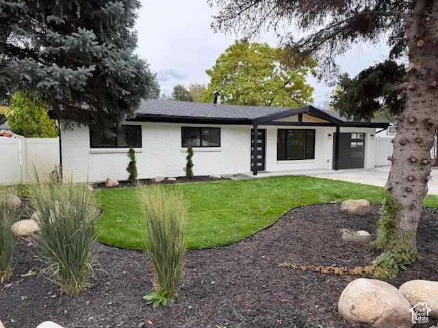 Single-level home exterior featuring painted brick masonry, dark-trimmed window frames, an integrated garage with modern door, and landscaped front yard with mulch beds and mature coniferous trees