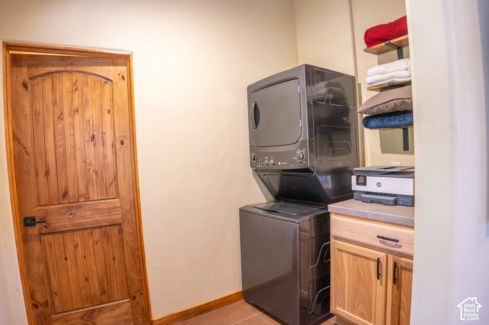 Laundry area featuring a knotty wood-finish arched door with matte black hardware, stacked metallic laundry appliances, custom light-wood base cabinetry with stone-finish countertop, and industrial-style wall-mounted shelving