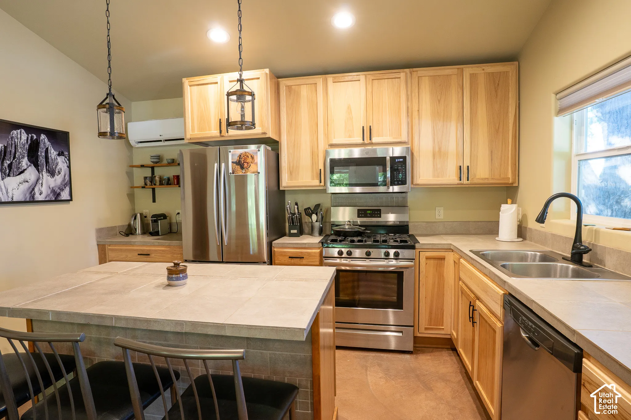 Kitchen featuring natural wood cabinetry, stainless steel appliances, tiled island countertop, double-basin sink with matte black faucet, and pendant lighting